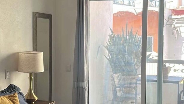 Une chambre d’hôtel équipée d’un lit, d’un ventilateur de plafond, d’une lampe, d’un miroir et d’une porte-fenêtre coulissante donnant sur un espace extérieur.