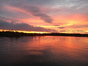 Un coucher de soleil sur une étendue d’eau calme, avec la silhouette d’arbres et un ciel nuageux.