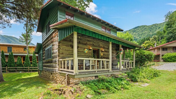 A rustic log cabin with a porch, stone foundation, and a mountain backdrop. - MOUNTAIN JOY COTTAGES #5 - STARS AND STRIPES (Maggie Valley)