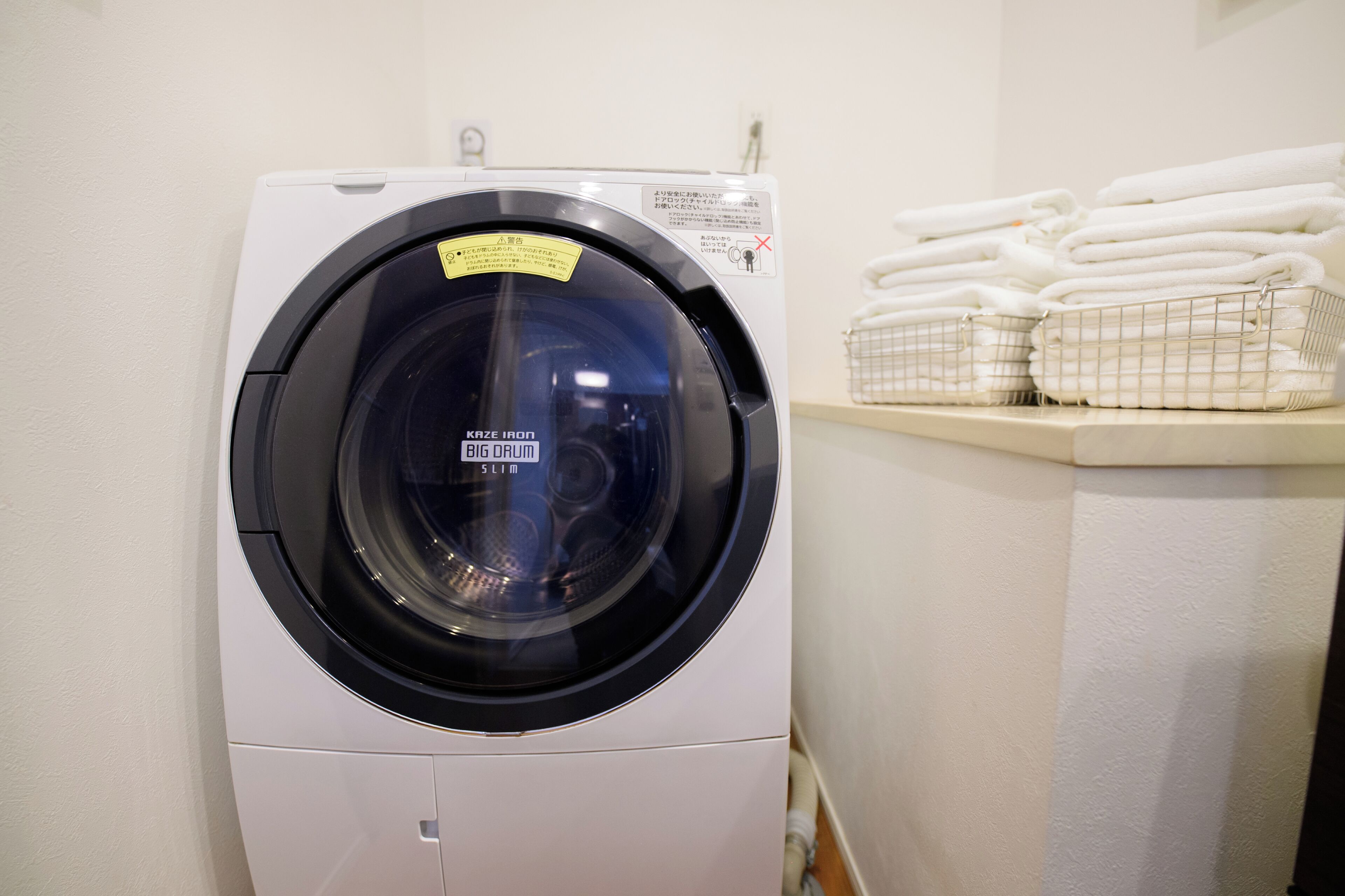 A front-loading washing machine with a label, stacked towels, and a metal laundry basket.