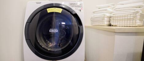 A front-loading washing machine with a label, stacked towels, and a metal laundry basket.