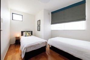 Une chambre avec deux lits simples, une table de chevet avec une lampe, une fenêtre avec un store et un petit cadre photo accroché au mur.