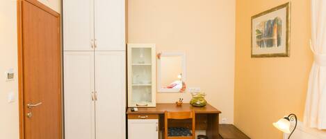 Habitación pequeña con un armario de madera, un escritorio con silla, un refrigerador compacto, un cesto de basura y una mesita de noche con lámpara.