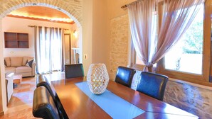 A dining area with a wooden table, chairs, and a decorative vase. There is a stone wall and a window with curtains.