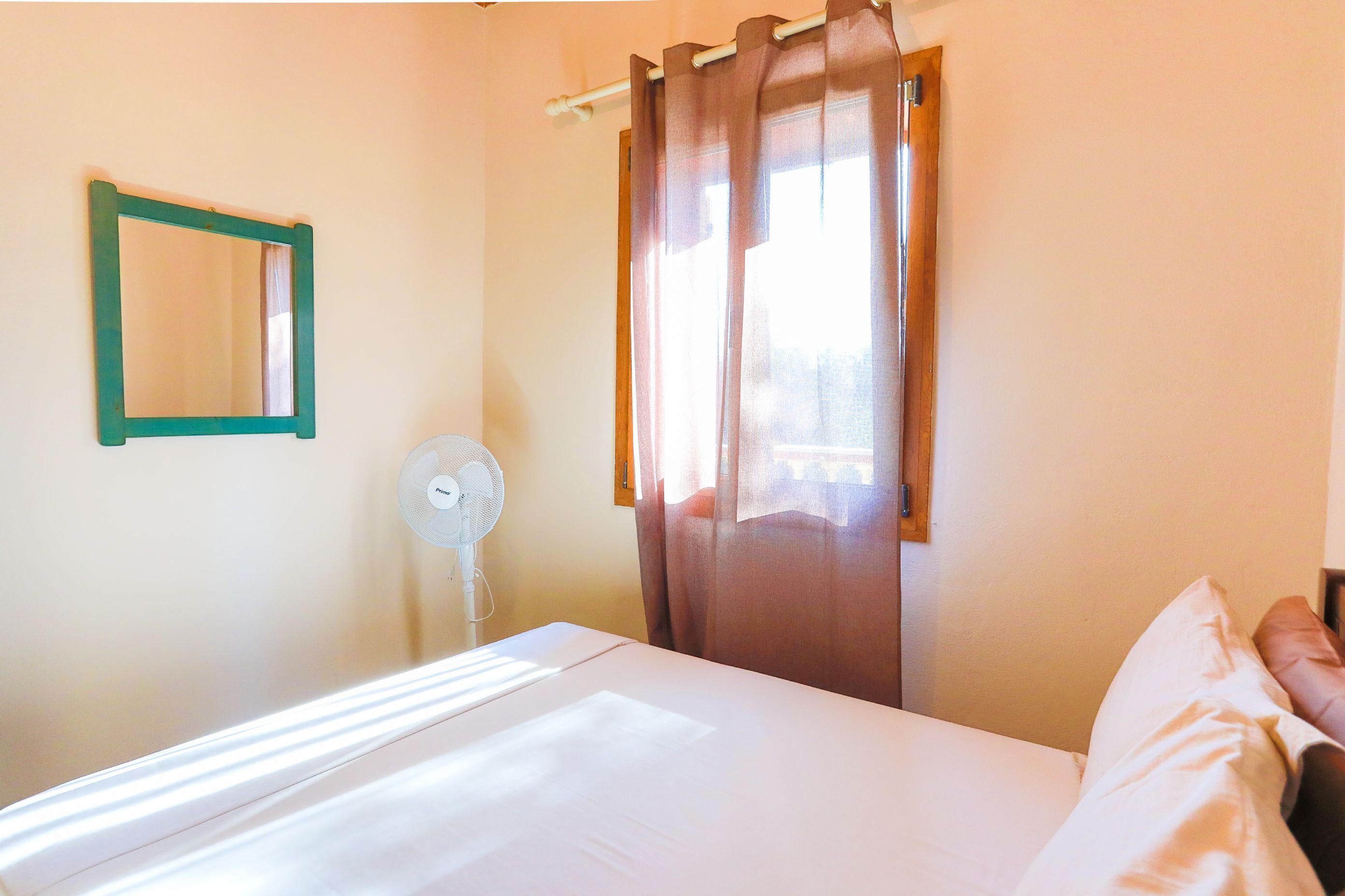 Quarto com cama, ventilador, janela com cortinas e espelho fixado na parede.