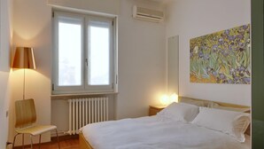 Ein Schlafzimmer mit Bett, Stuhl, Fenster und Klimaanlage. Ein Gemälde hängt an der Wand.