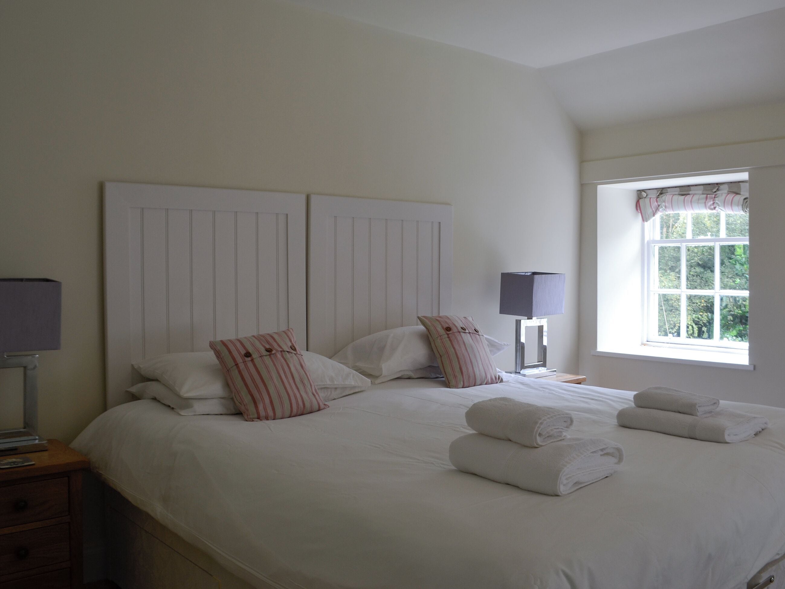 Una cama bien hecha con sábanas blancas y almohadas a rayas, una lámpara de noche y una ventana con cortinas.