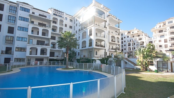 A multi-story residential building with balconies, a swimming pool, and palm trees.