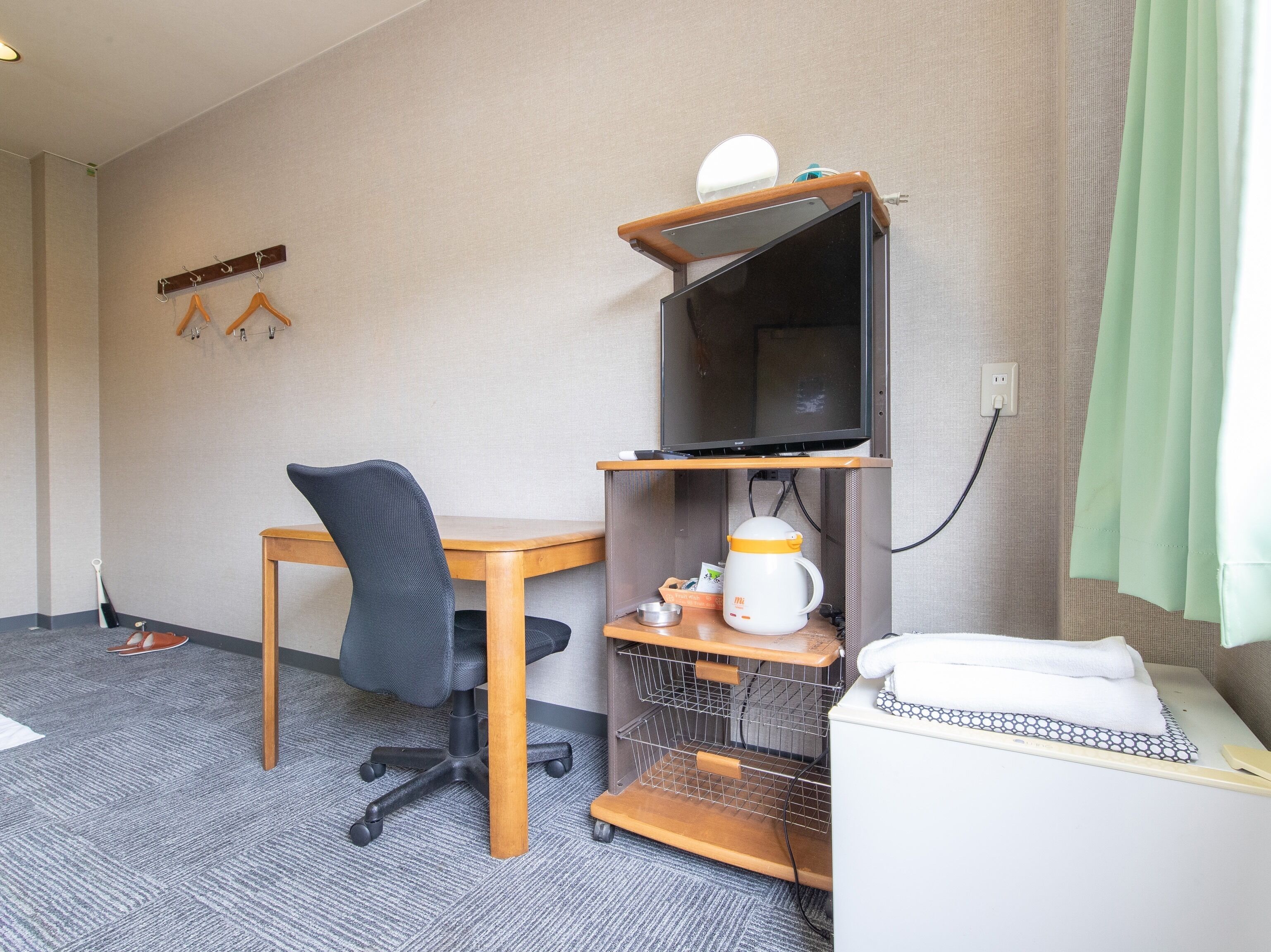 A hotel room with a wooden desk, a black office chair, a television on a stand, a kettle, a small shelf with toiletries, and a bed with a white blanket.