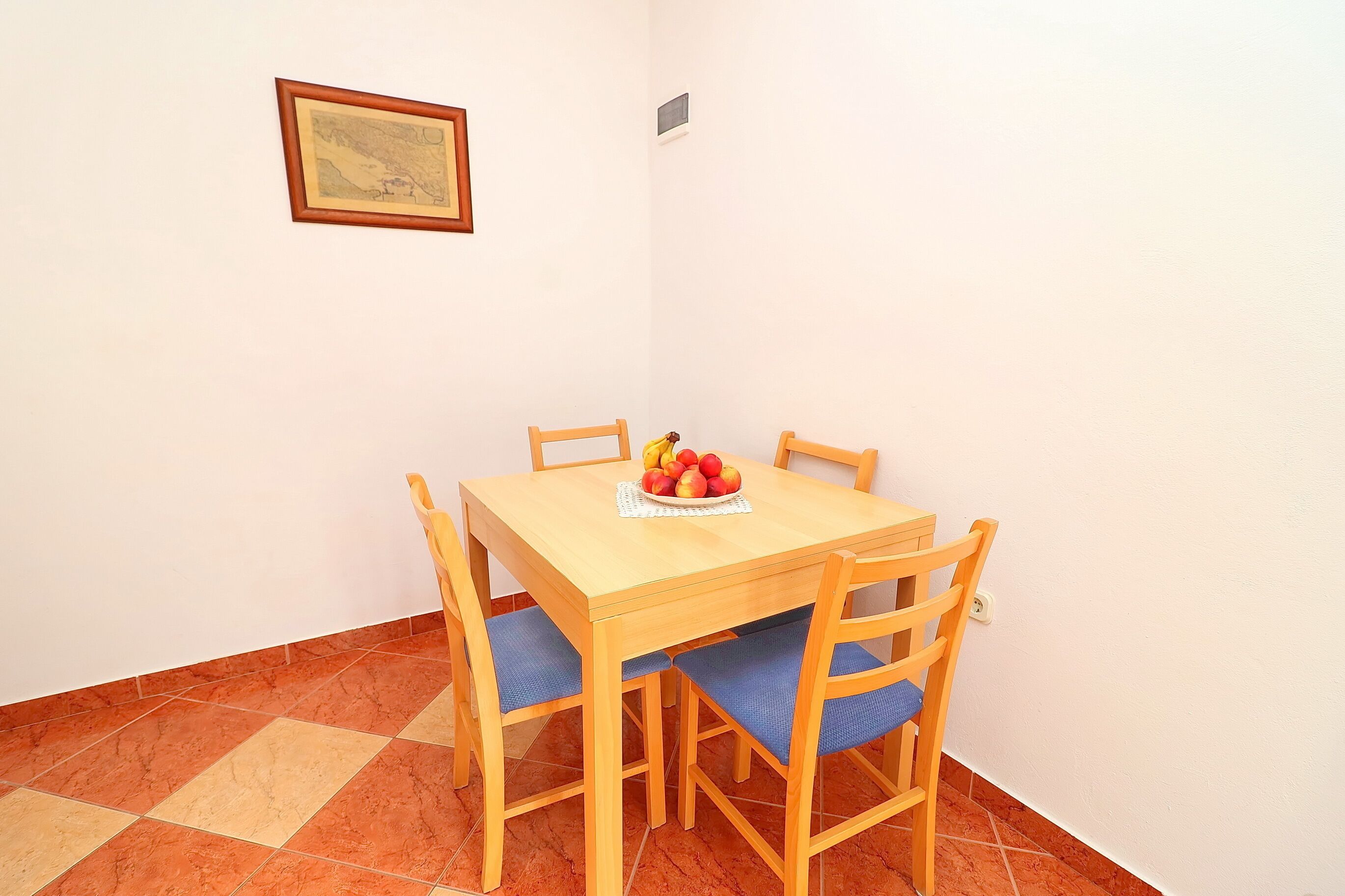 Ein Essbereich mit einem Holztisch und Stühlen, einer kleinen Obstschale auf dem Tisch und einem gerahmten Bild an der Wand.