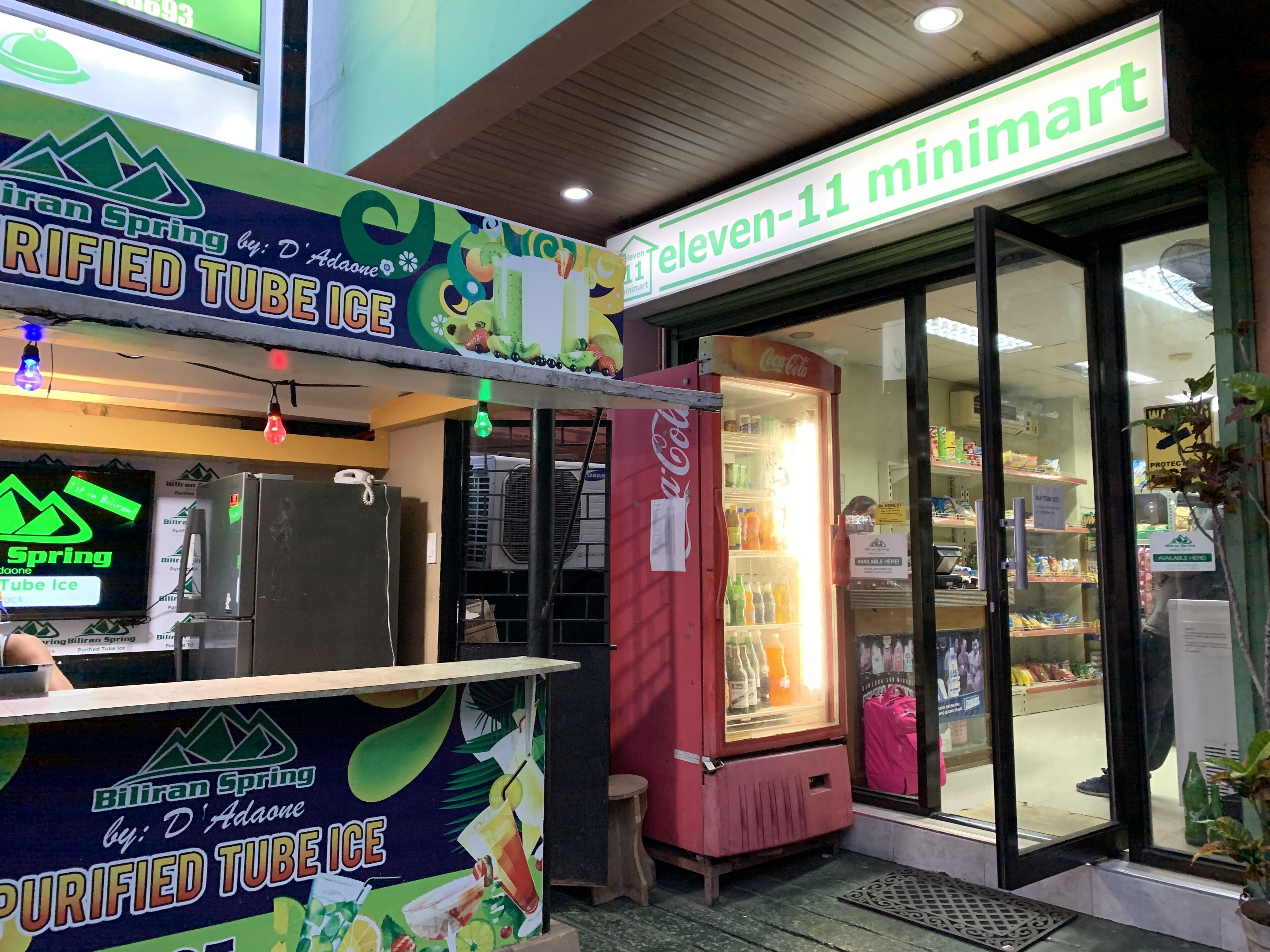 A small outdoor kiosk with a sign for 'Biliran Spring by D' Adaone' and a Coca-Cola refrigerator.