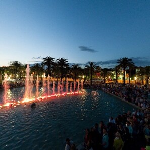 Ein Außenpool bei Nacht mit Springbrunnen, umgeben von Palmen und vielen Menschen.