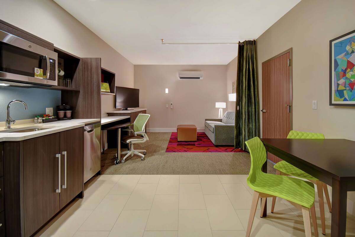 A compact hotel room with a kitchenette, a dining area, a sofa, and a TV.