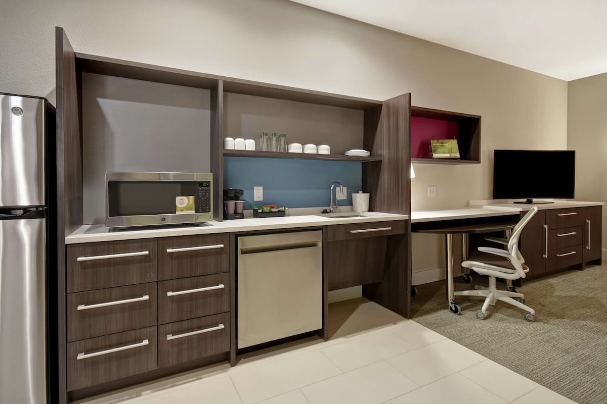 A modern kitchen with a built-in microwave, stainless steel refrigerator, and a dishwasher. There is a sink with a faucet, a countertop with various items, and a television mounted on the wall.