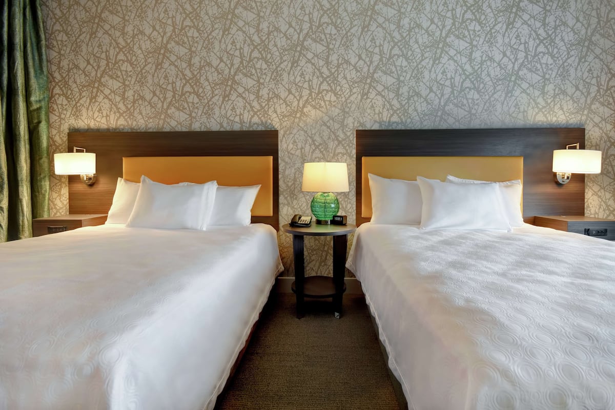 A hotel room with two beds, a small table with a lamp, and a patterned wallpaper.