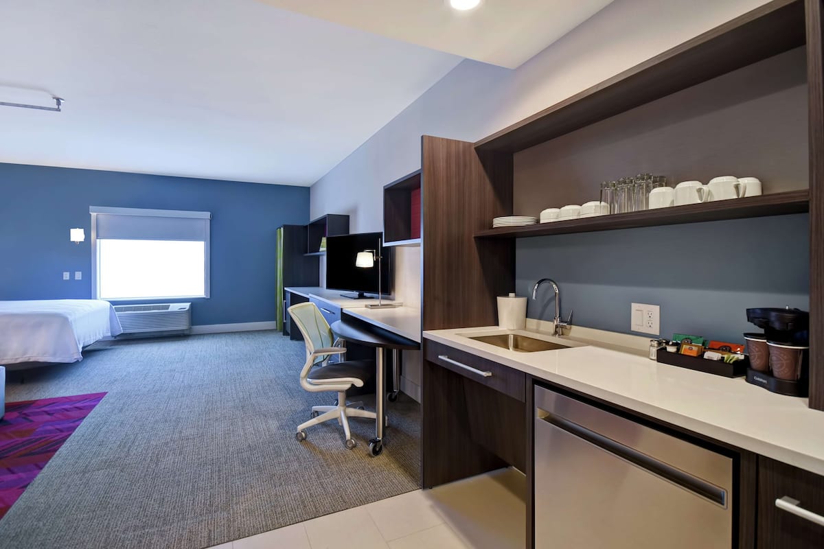 A modern hotel room with a bed, desk, chair, kitchenette, and a large window.