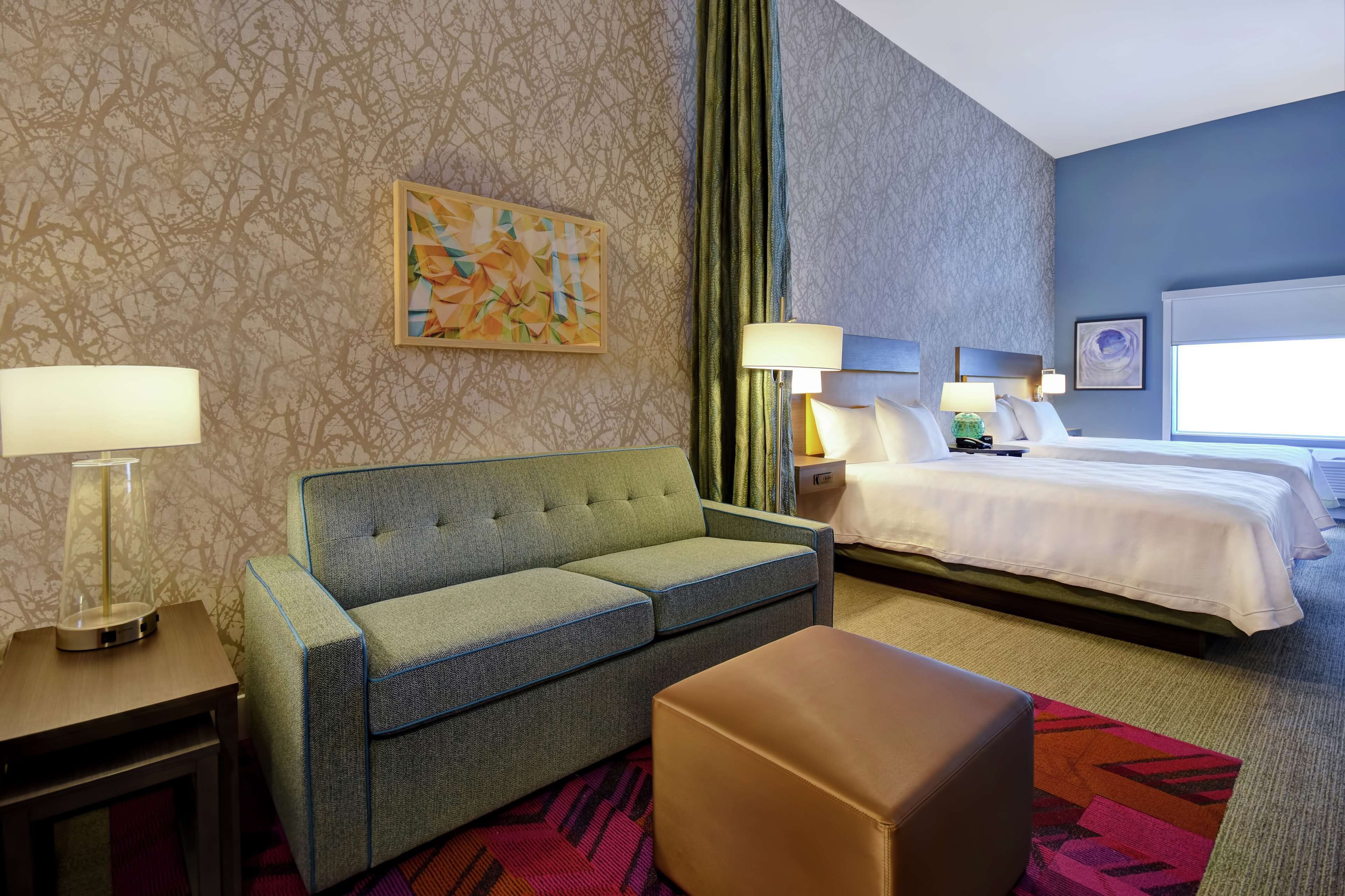 Ein Hotelzimmer mit einem grünen Sofa, einem Bett mit weißen Laken, einem Nachttisch mit Lampe und einem farbenfrohen Teppich.