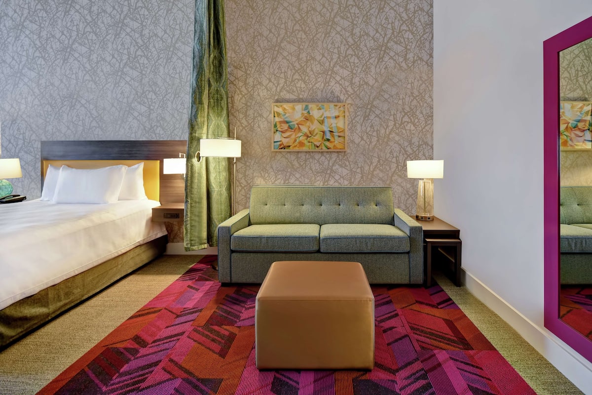 A hotel room with a bed, a sofa, an ottoman, and two nightstands with lamps.