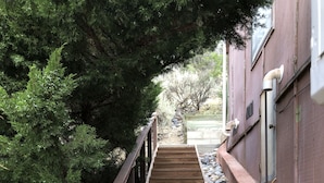 A wooden staircase with a metal railing, surrounded by rocks and greenery, leading to a building with a window and a utility box. - High Desert Perch with views. (Farmington)