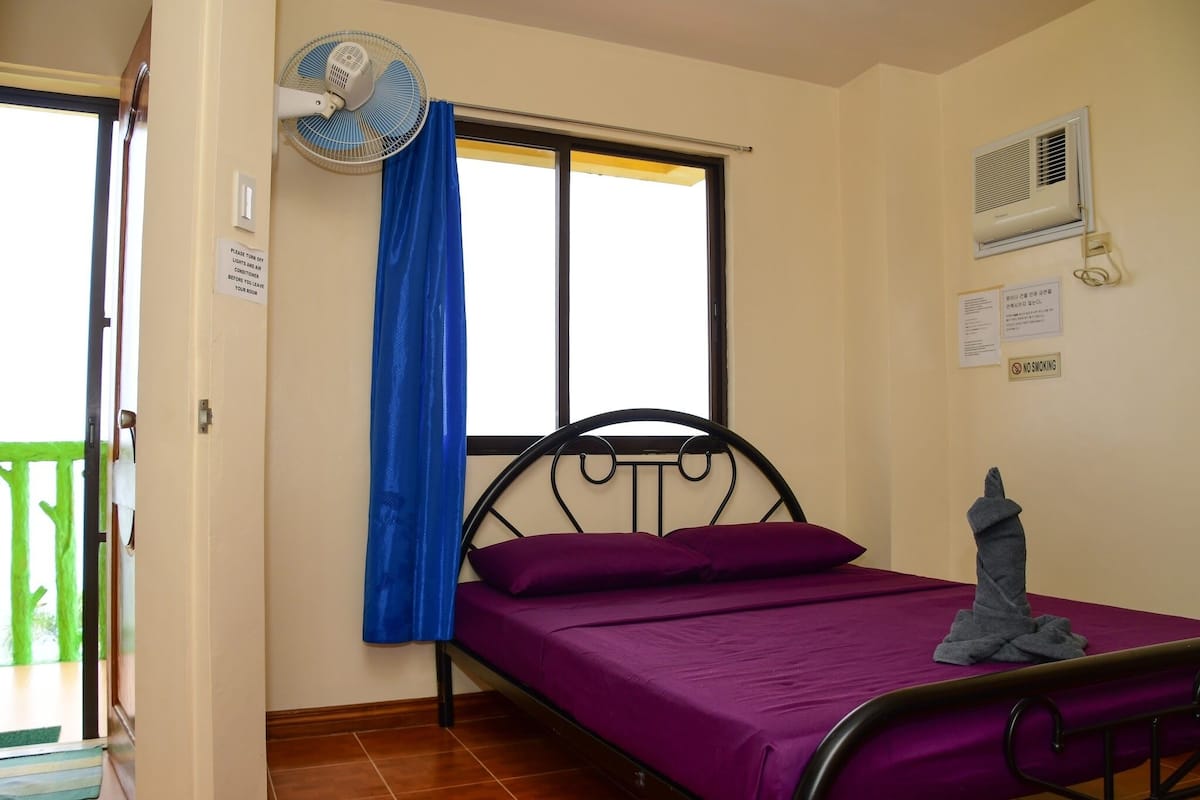 A single bed with a purple mattress, a blue curtain, a wall-mounted fan, and a no-smoking sign.