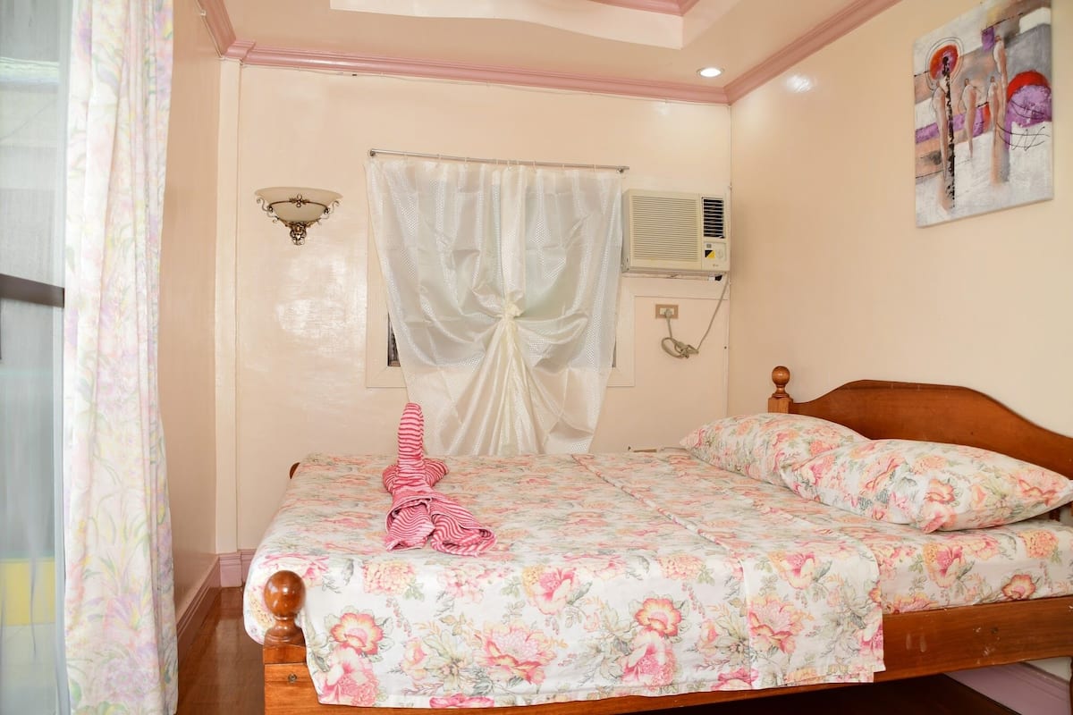 A bedroom with a wooden bed, floral bedding, a white curtain, and a wall-mounted air conditioner.