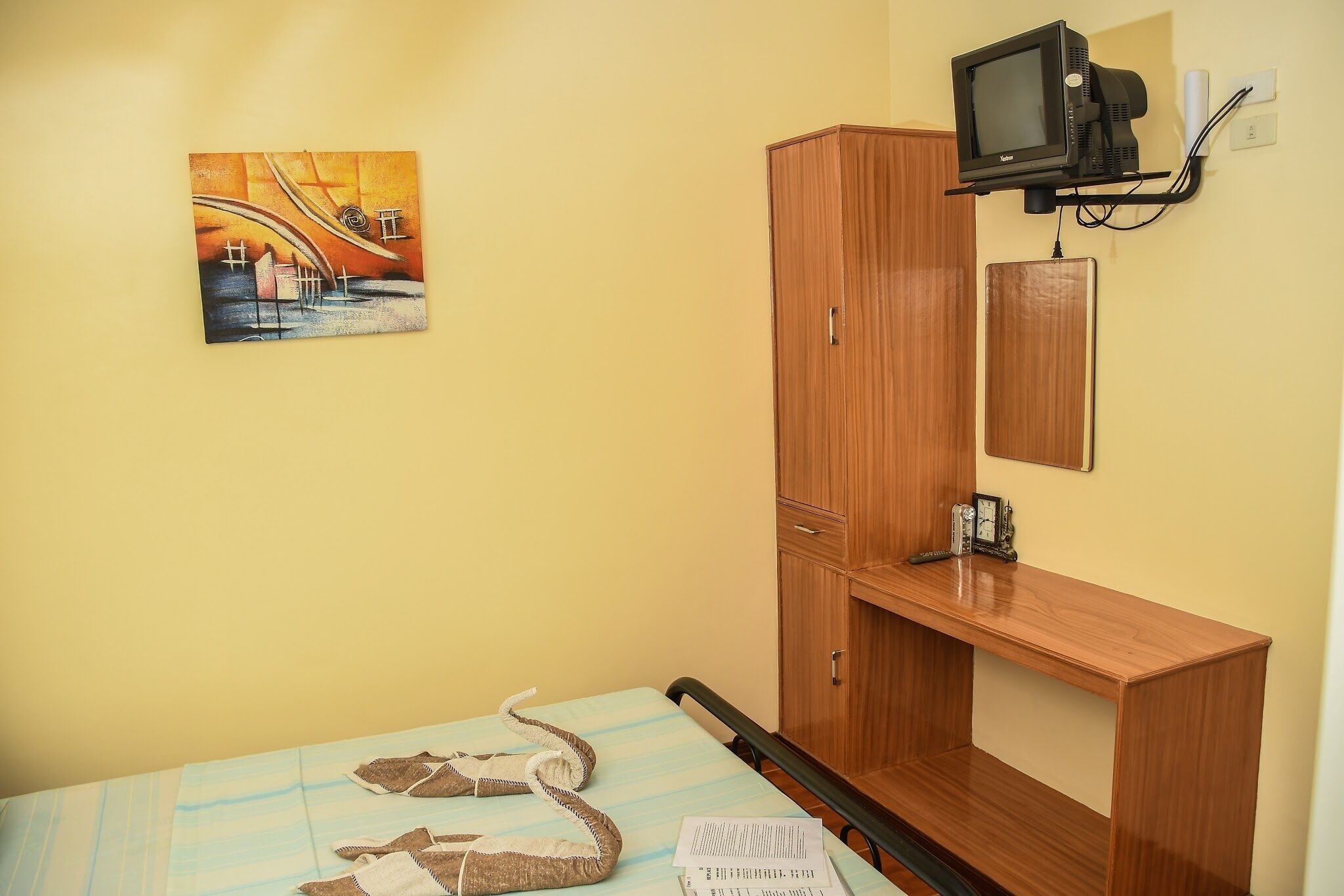 A room with a bed, a wooden desk, a television mounted on the wall, and a painting on the wall.