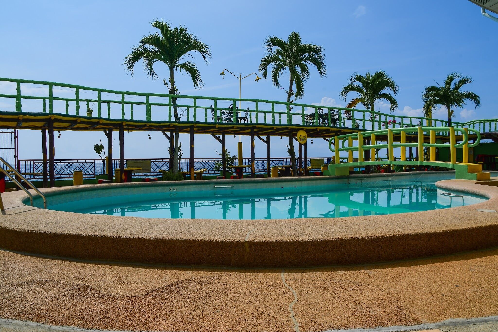 Une piscine bordée d’une terrasse verte et jaune, des palmiers et un ciel d’un bleu limpide.