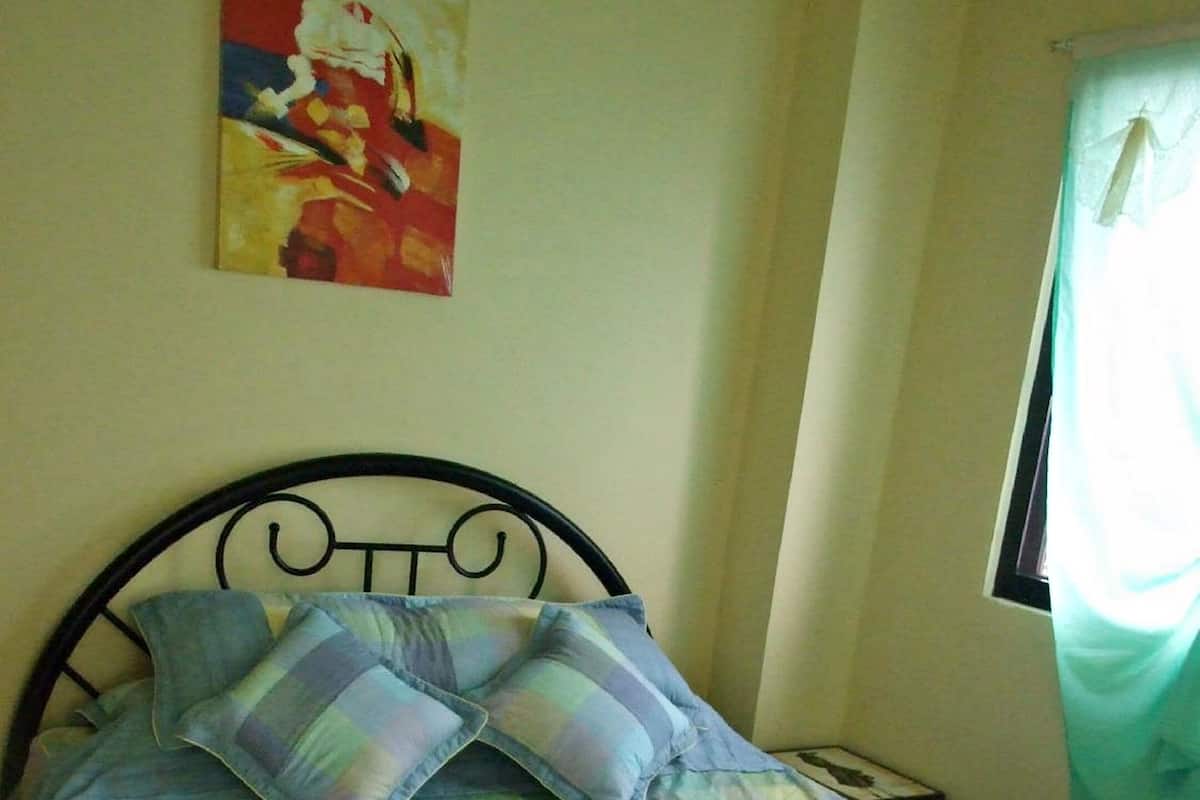 A bed with a checkered bedspread and two pillows, a metal headboard, a small bedside table, and a painting on the wall.
