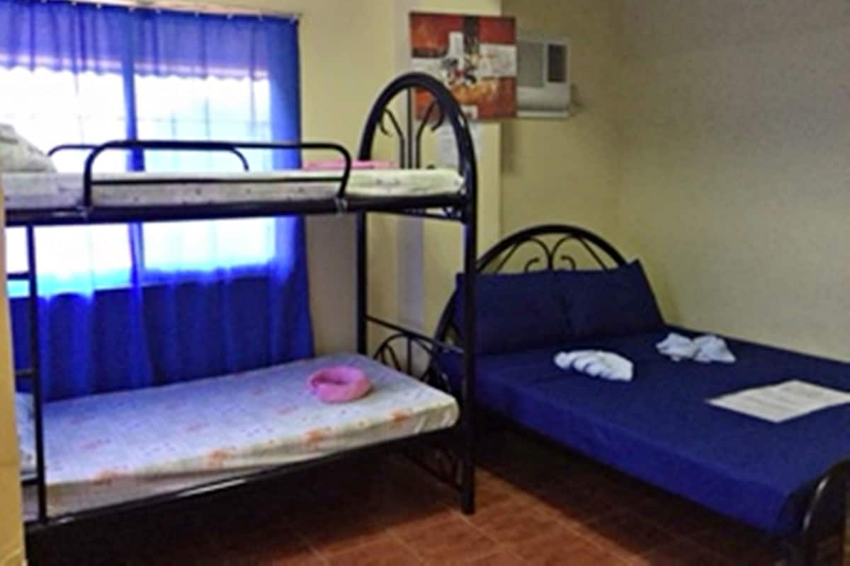 A dormitory room with bunk beds, a single bed, a window with blue curtains, and a wall-mounted air conditioner.