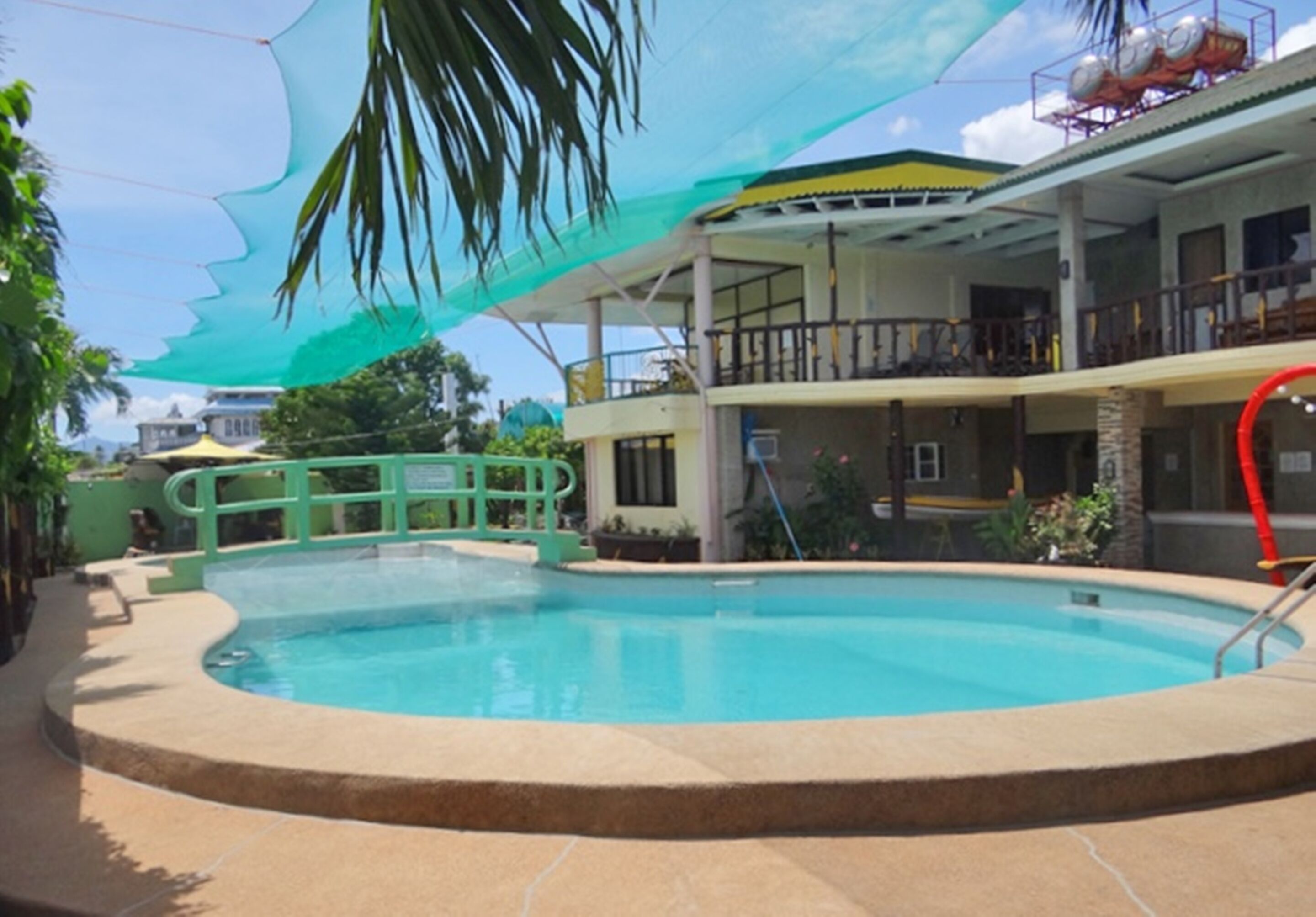 Una piscina con borde curvo, rodeada por un solado de hormigón. Se observa un dosel verde sobre ella, y un edificio con balcones al fondo.