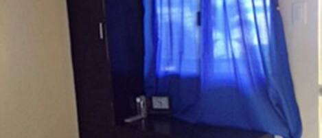 A small room with a bed, a desk, a television mounted on the wall, and a window with blue curtains.