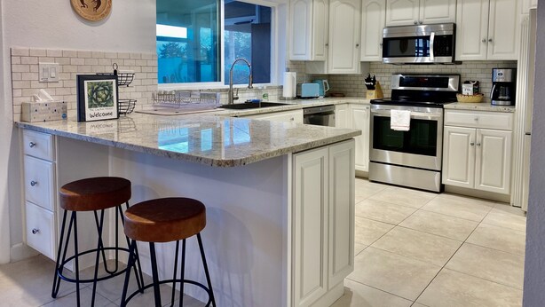 A modern kitchen with white cabinets, a stainless steel oven, and a microwave. There is a large island with stools and a framed welcome sign. - Cactus House - Spacious home & pool next to mountains and close to everything (Phoenix)