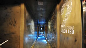 A narrow hallway with a blue-lit exit sign, a door labeled 'EMERYSTUDIO/NO COLOR LED', and a wall with various text and graphics. - 7 Motel (Gimhae)