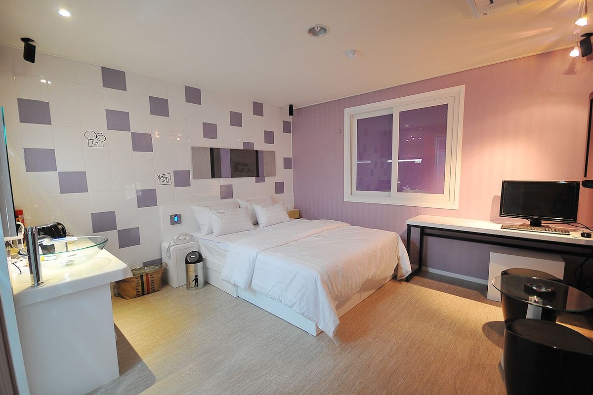 A modern hotel room with a bed, bedside table, desk, and computer.