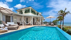 A poolside area with a clear view of the ocean, a covered seating area with lounge chairs, and a building with a balcony overlooking the pool.