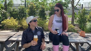 Deux personnes sont assises à une table en bois à l’extérieur, l’une tenant une bouteille de bière et l’autre une canette.