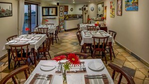 A dining area with multiple tables set for meals, a kitchen in the background, and artwork on the walls. - Quiet guest room with double bed and kitchen, near Port Elizabeth (Port Elizabeth)