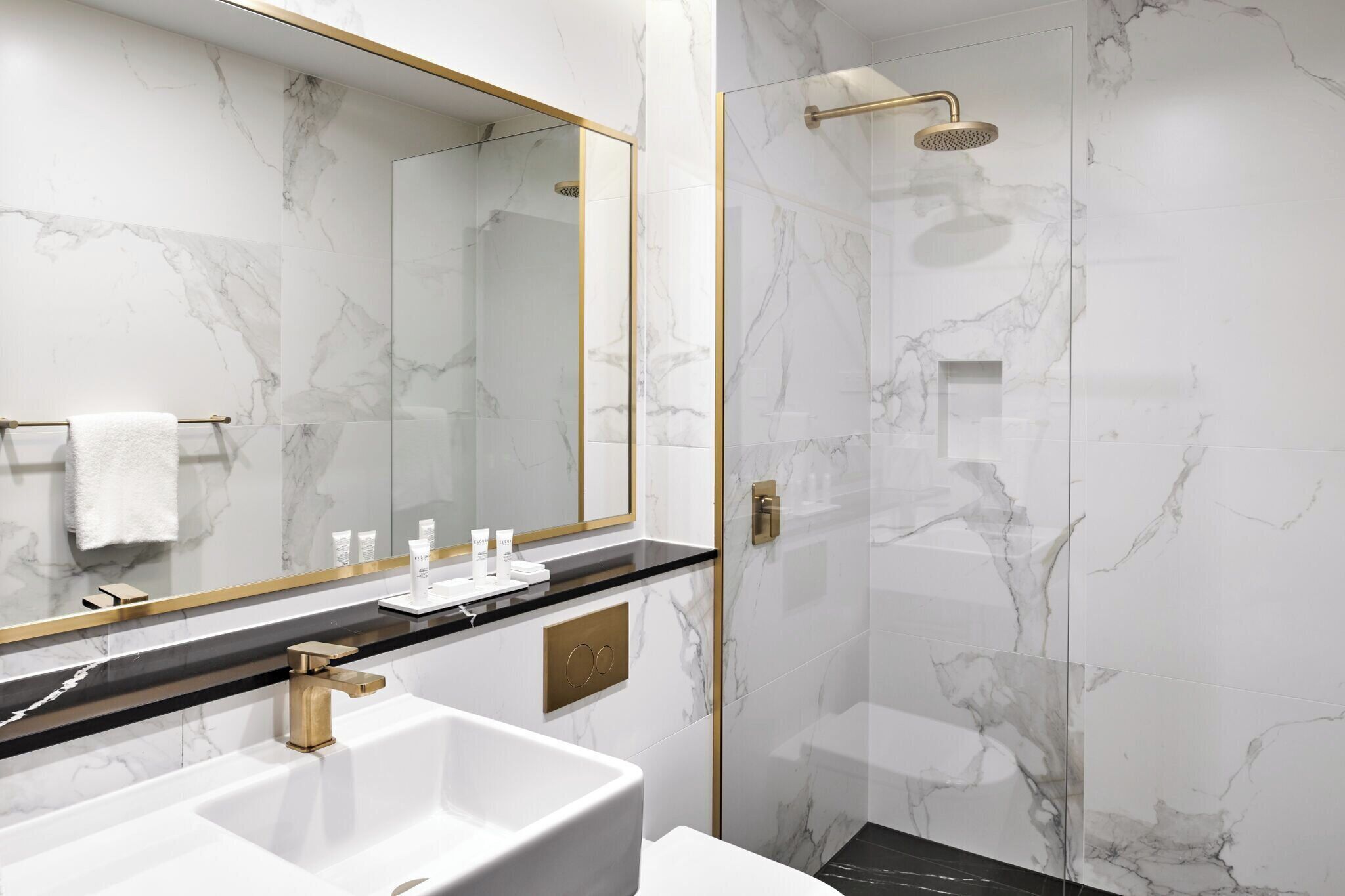 Un baño moderno con pared de mármol, un espejo grande, un grifo dorado y una ducha con cabezal de ducha dorado.