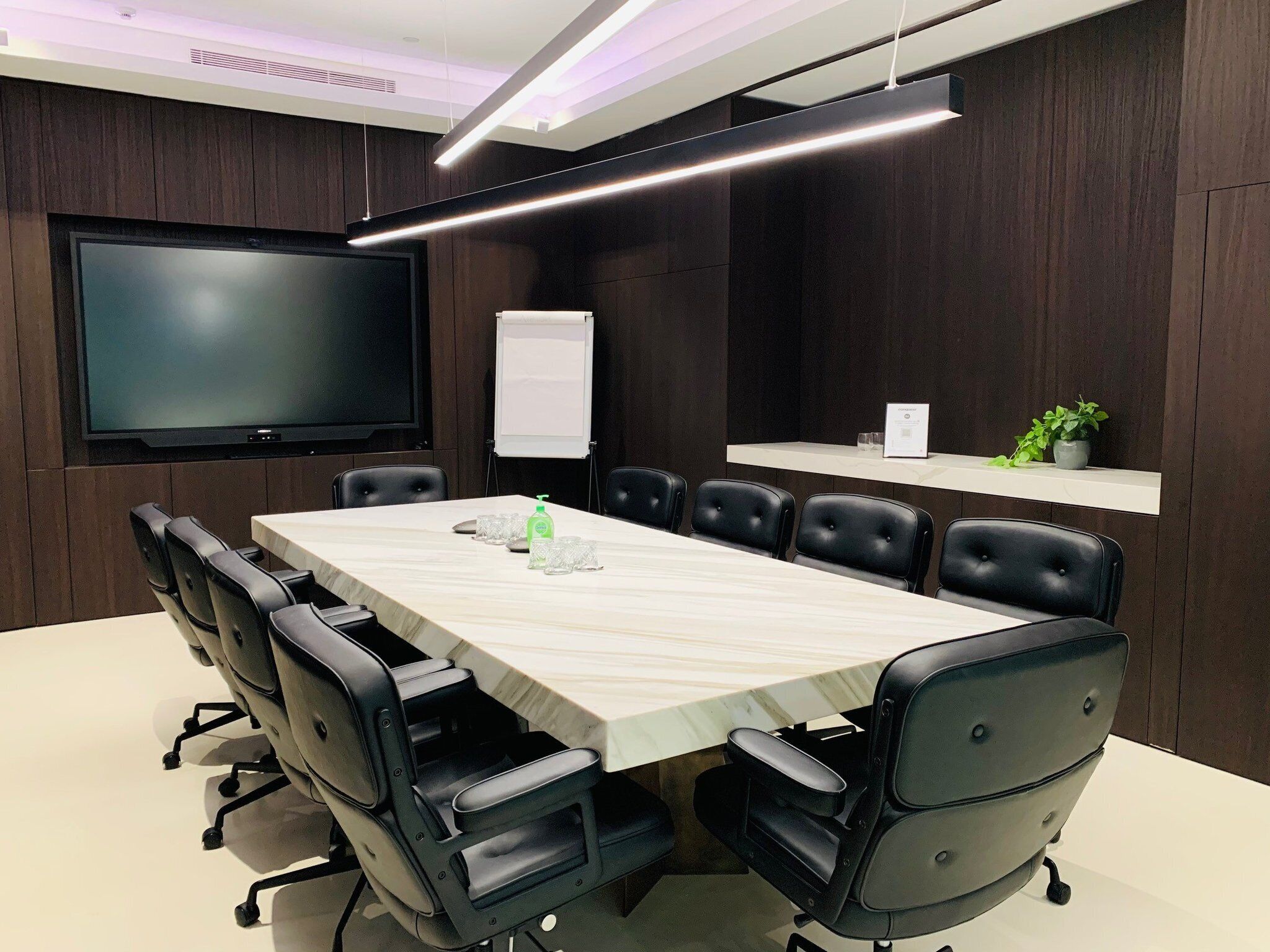 A conference room with a long table, black chairs, a flat-screen TV, a whiteboard, and a shelf with a plant.