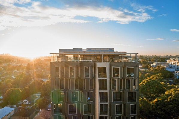 A modern high-rise building with multiple balconies and large windows, surrounded by greenery and other residential buildings.