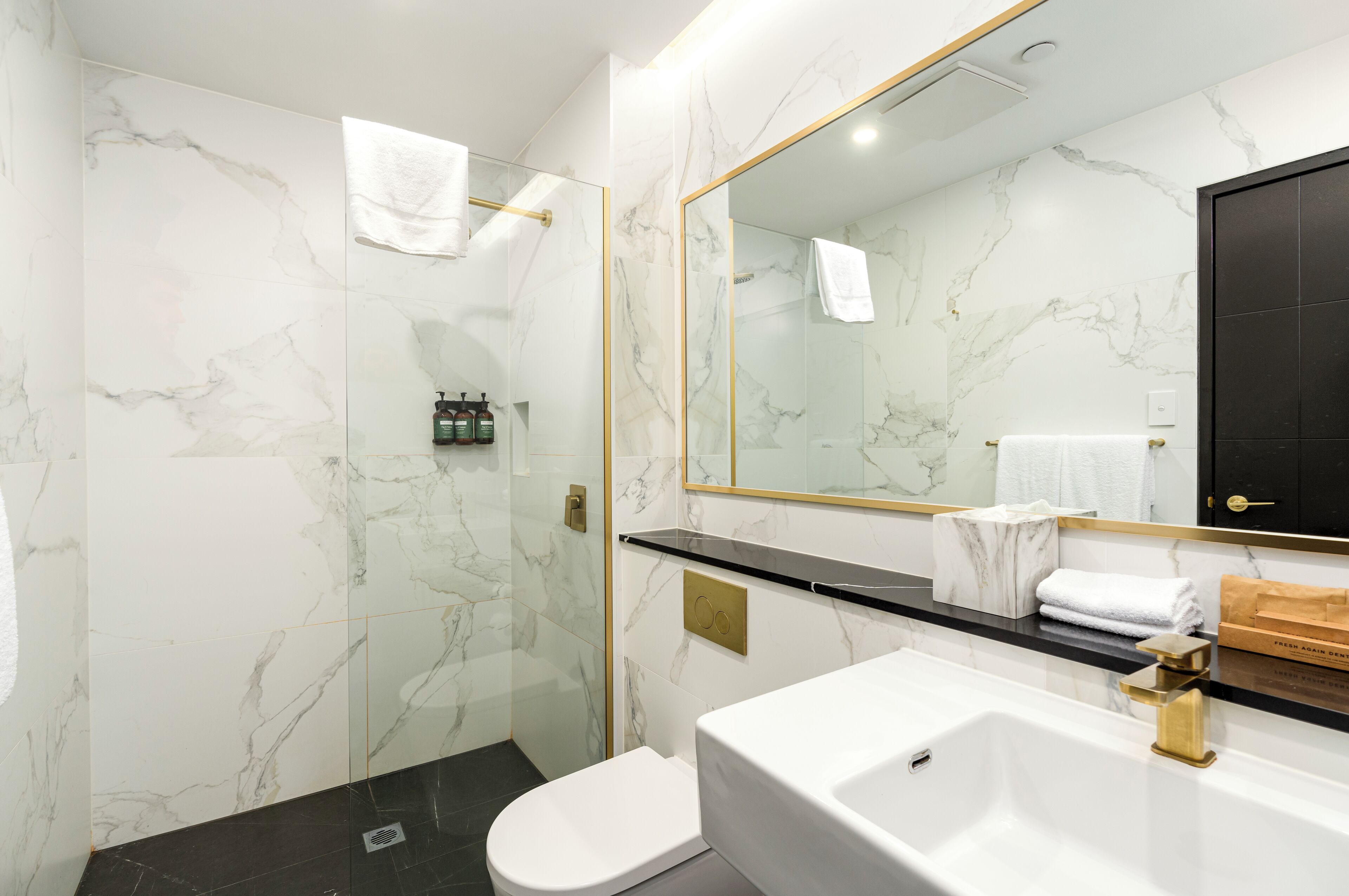 A modern bathroom with a marble wall, a glass shower enclosure, a white toilet, a white sink with a gold faucet, and a large mirror.