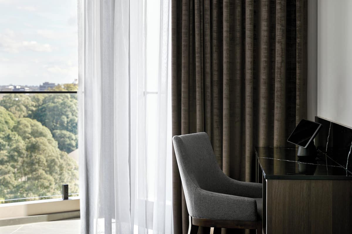 A modern room with a view of a forested area, a grey armchair, and a black desk with a laptop.