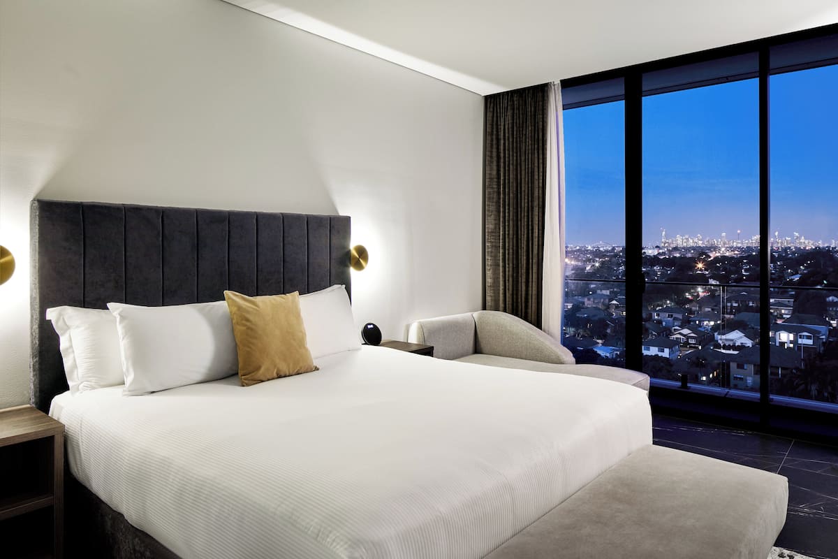 A modern hotel room with a large bed, a view of the city at dusk, and a large window.