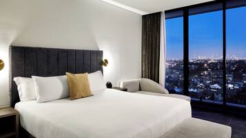 A modern hotel room with a large bed, a view of a cityscape through a large window, and a nightstand with a lamp.