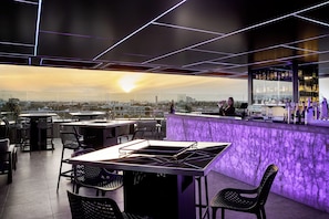 Un espace bar extérieur avec vue sur la ville, comprenant des tables, des chaises et un barman.
