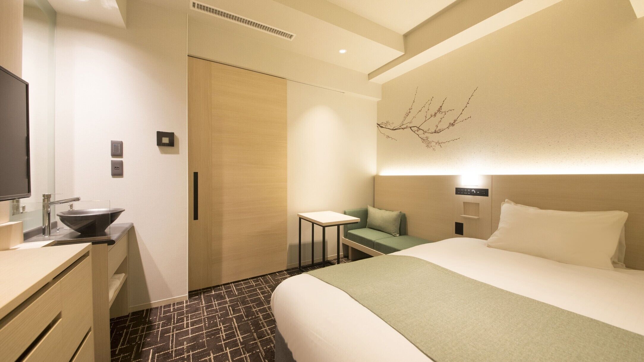 Une chambre d’hôtel comprenant un lit, une table de chevet, une petite chaise, un bureau, un lavabo et un miroir.