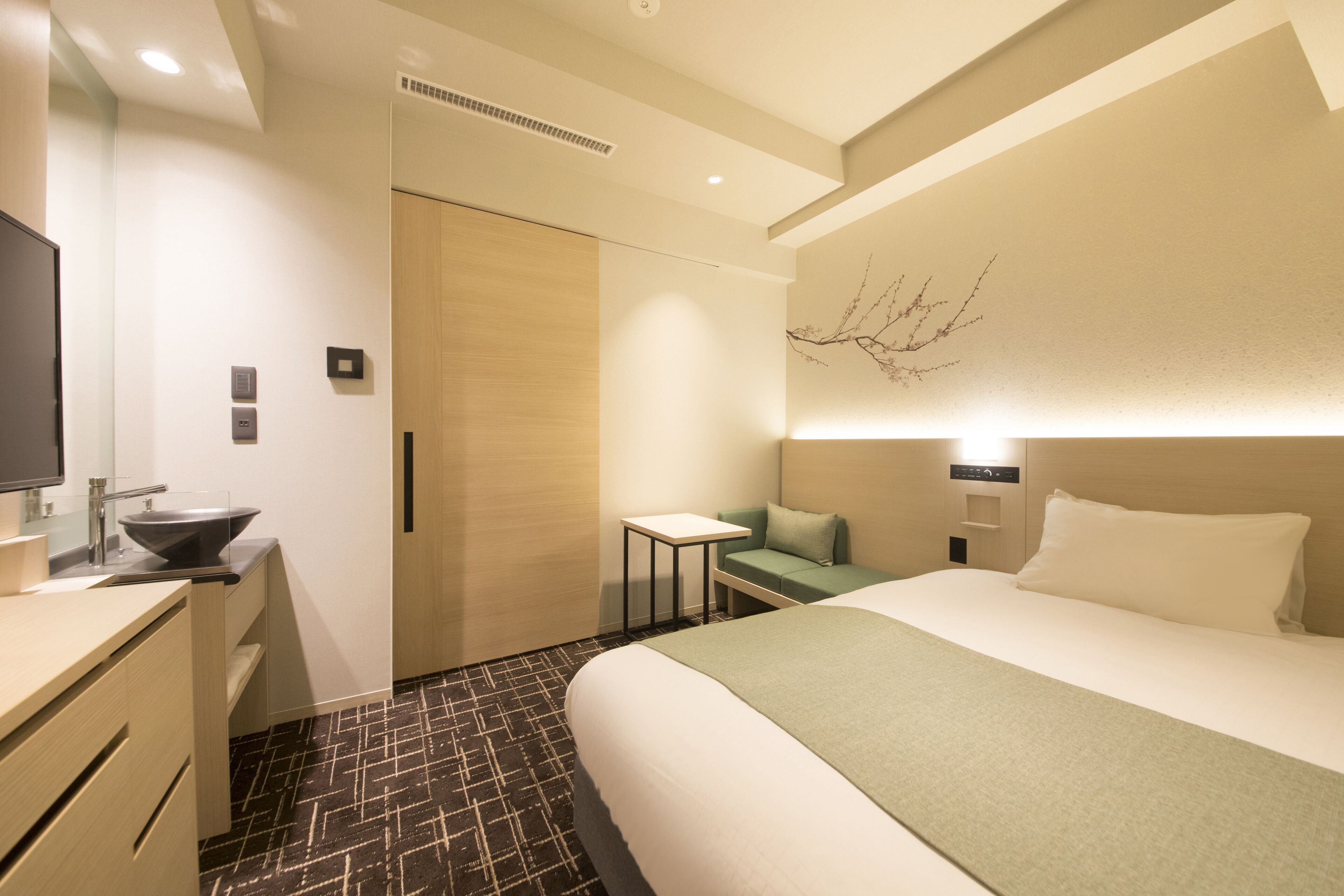 A hotel room with a bed, a desk with a sink, a small table, and a wall-mounted TV.