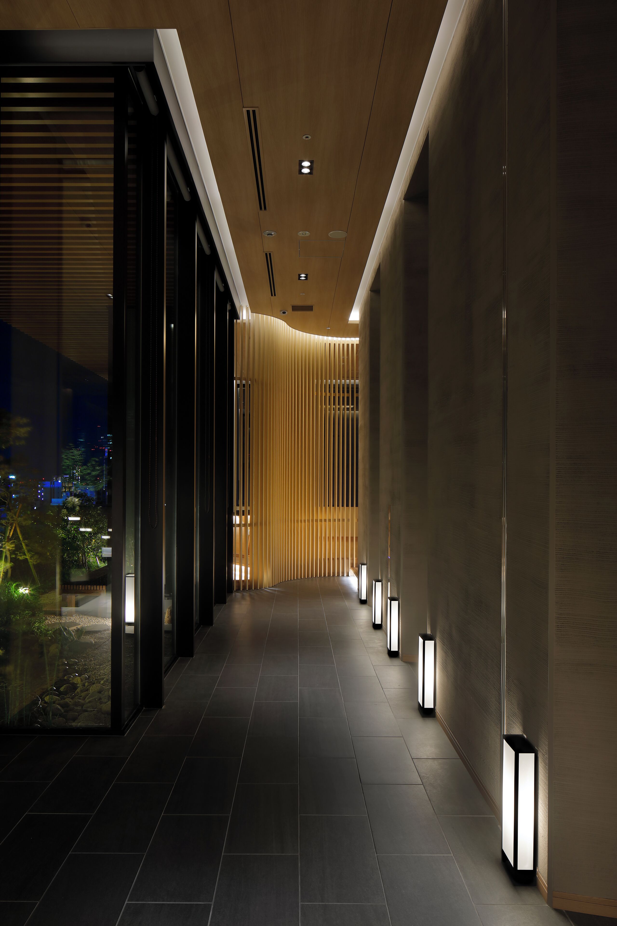 A modern hallway with a wooden slat wall, recessed lighting, and a view of the cityscape through large windows.