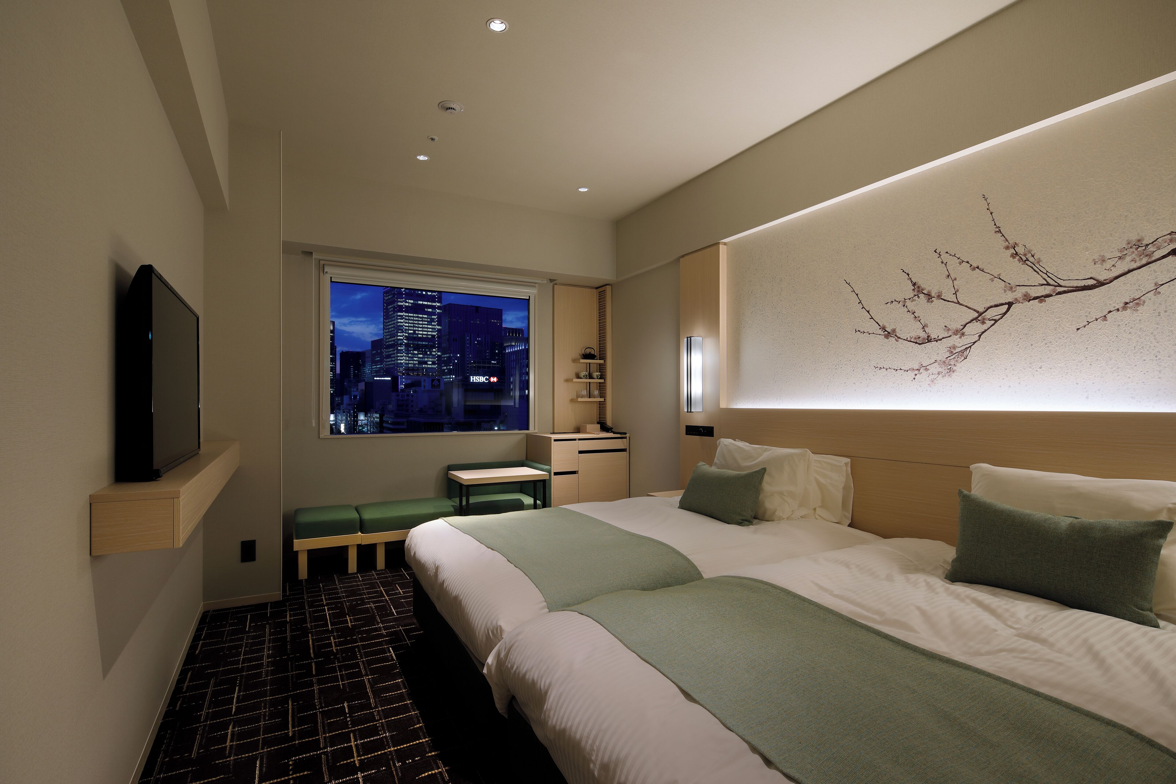 A modern hotel room with a large bed, a desk, a bench, and a city view.