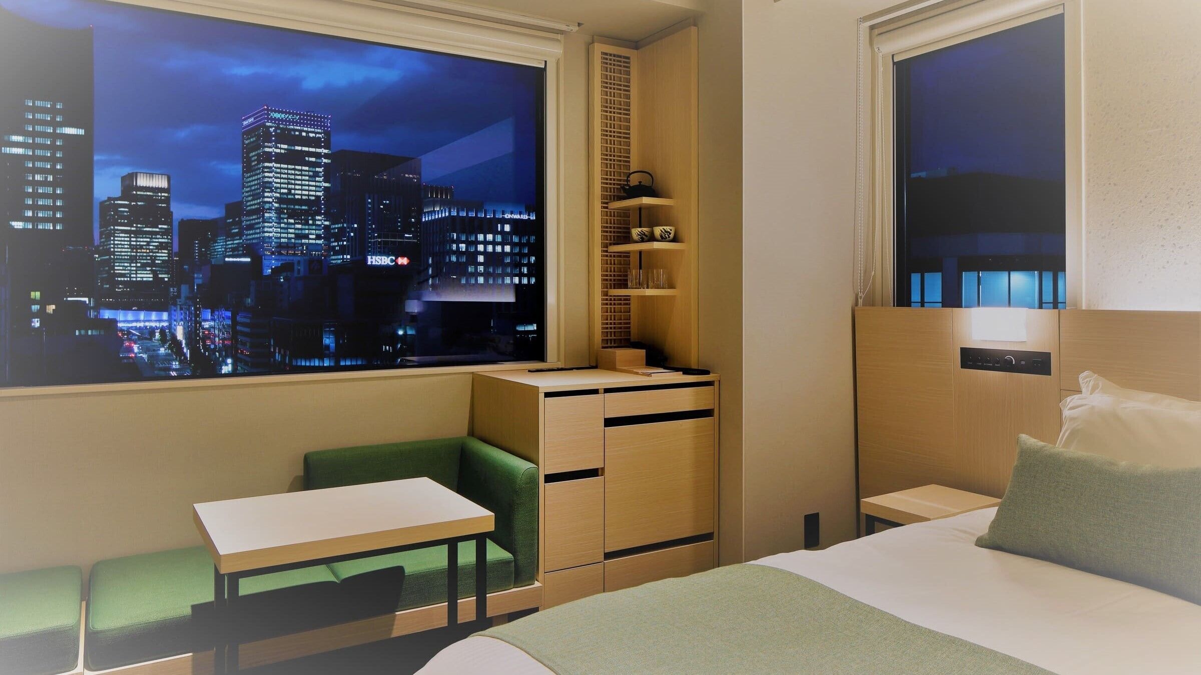 Une chambre d’hôtel avec vue sur la ville, un bureau, une chaise, un lit et une petite étagère avec quelques objets.